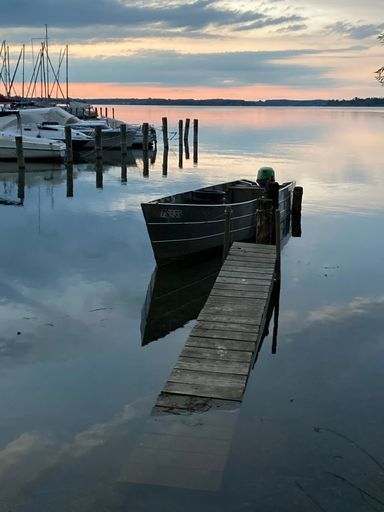 A serene lakeside scene with boats moored at a wooden dock during sunset, reflecting on calm waters.