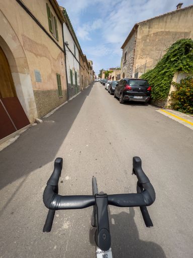 Cycling down a narrow street lined with rustic buildings and parked cars under a partly cloudy sky.