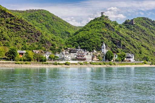 Scenic view of a quaint village along the Rhine River, surrounded by lush green hills and a historic castle, under a partly cloudy sky.