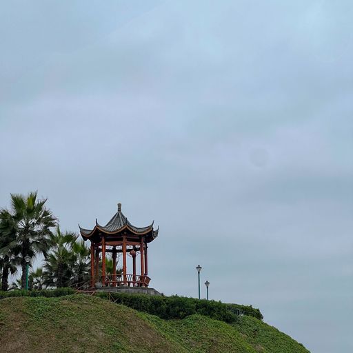 A traditional Asian gazebo on a grassy hilltop under a cloudy sky, surrounded by palm trees.