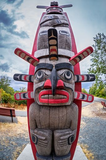 A vibrant totem pole stands tall against a cloudy sky in a park setting, showcasing intricate carvings and vivid colors.
