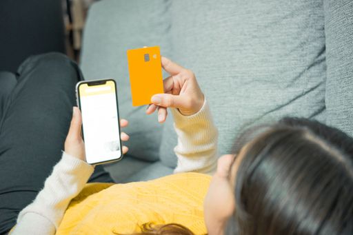 A young woman in casual attire shops online using a smartphone and an orange credit card while relaxing on a gray sofa.