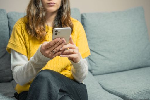 Young woman in a yellow sweater using a smartphone on a gray sofa, focusing on the device and her hands.
