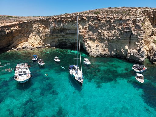 Boats and swimmers enjoy the crystal-clear turquoise waters by the rocky cliffs of Blue Lagoon, Malta, under a bright blue sky.