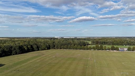 Aerial view of a vast sports field with soccer pitches, surrounded by lush greenery under a partly cloudy sky.