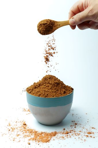 Cocoa powder falling from a wooden spoon into a bowl on a white background, creating a cloud of dust