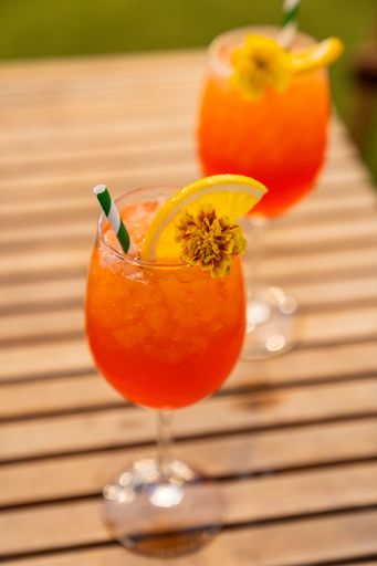 Close-up of an Aperol Spritz served in a wine glass with crushed ice, lemon slice, edible flower and paper straw, on a wooden table outdoors.