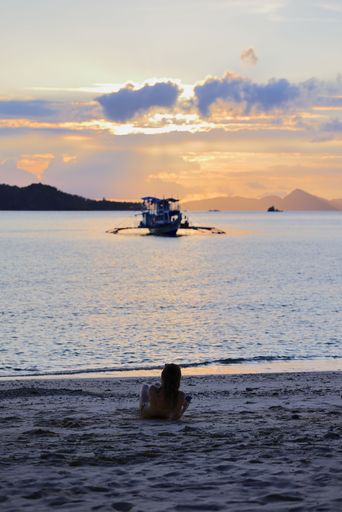A serene beach scene at sunset with a woman relaxing on the sand, a boat on the water, and a vibrant sky.