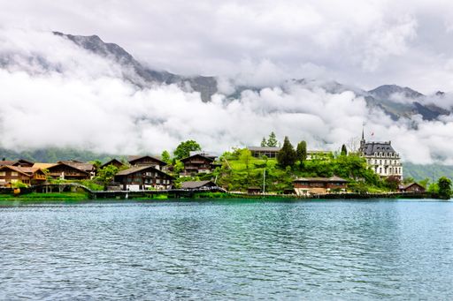 Scenic view of a Swiss lakeside village with charming houses and a castle, surrounded by misty mountains and lush greenery.