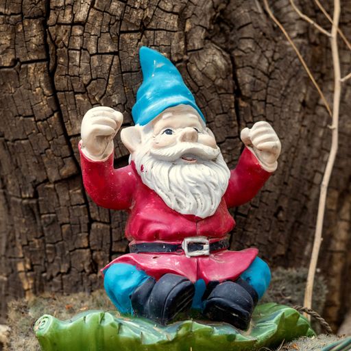 A colorful garden gnome with a red jacket and blue hat sits cheerfully in front of a weathered wooden tree trunk.