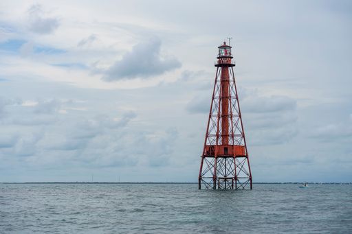 Red lighthouse tower standing in the ocean under a cloudy sky, with distant shoreline in the background.