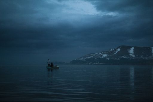 An eerie shot of two males in a small boat on the sea with mountain background in a gloomy day