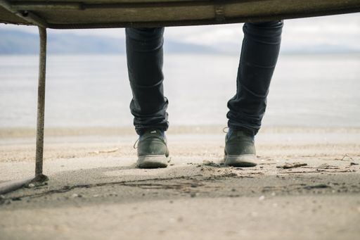 A low angle shot of dressed legs of a person sitting on a bench while stepping on a dirty sand