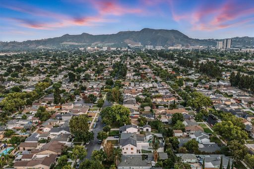 An aerial view of a suburban neighborhood with tree-lined streets and houses, under a dramatic, colorful sky at sunset.