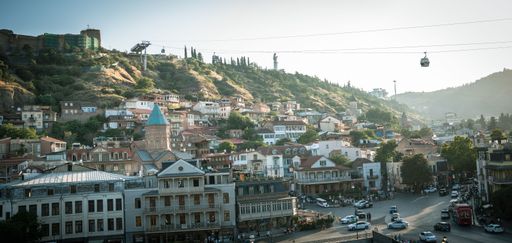 A wide view of Tbilisi, Georgia, featuring historic architecture, a bustling street, and a cable car against a hilly backdrop.