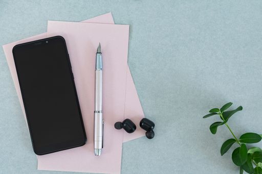 A top view of a smartphone next to a pink pen and black earbuds with a leaf on the side