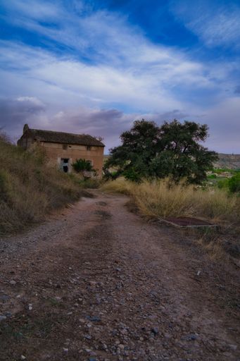A vertical shot of unpaved road leading to an old building under a  cloudy sky