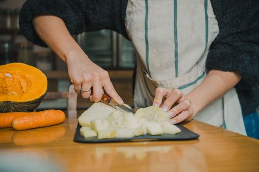 A person in apron slicing vegetables on a wooden table, including pumpkin and carrots, in a cozy kitchen setting.