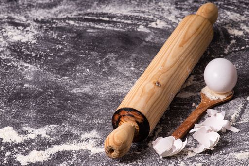 A closeup of the wooden rolling pin, spoon, and egg on the table covered with flour.