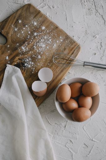 A vertical shot of a bowl of eggs and a silver whisk next to a chopping board on a white surface
