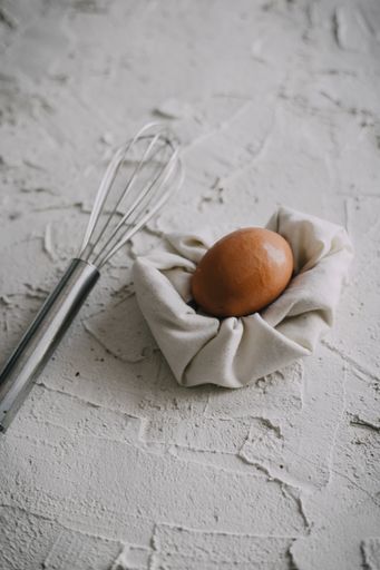 A hard-boiled egg on a white towel next to a silver whisk on a white surface