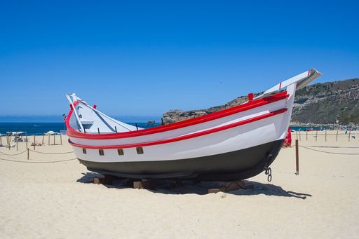 Nazare, Portugal, a traditional fishing boat with red and white details on a sandy beach, with the ocean and cliffs in the background.