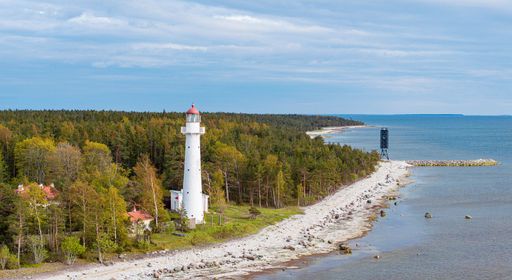 Scenic view of a coastal lighthouse surrounded by lush forest and rocky shoreline under a cloudy sky.
