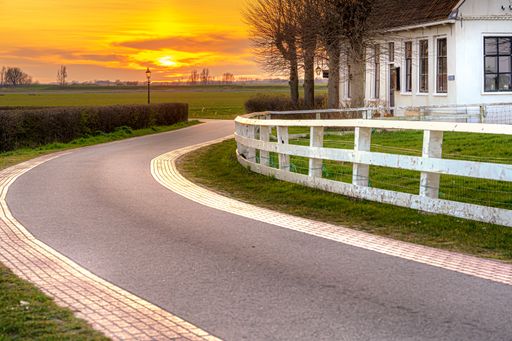 A beautiful shot of a concrete narrow road with a house near the field and a sunset in the background