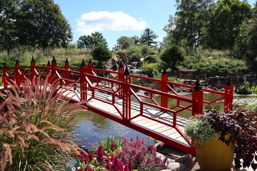 A vibrant red bridge over a serene pond in a lush garden landscape, under a clear blue sky.