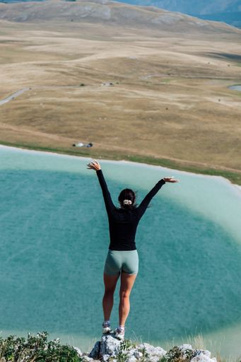 A young woman stands triumphantly on a cliff overlooking a serene lake with arms raised, surrounded by a vast landscape.