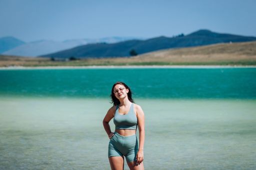 Young woman in sportswear standing by a turquoise lake with distant hills under a clear sky.