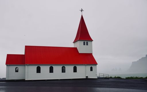 A picturesque church with a vivid red roof stands against a misty backdrop in Vik, Iceland, blending architecture with nature's serene ambiance.
