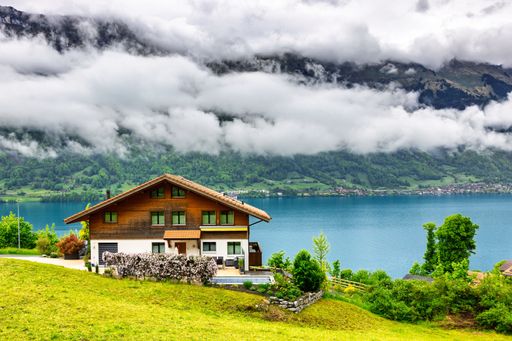 Scenic view of a Swiss chalet by a serene lake, surrounded by lush greenery and mountains under a cloudy sky.
