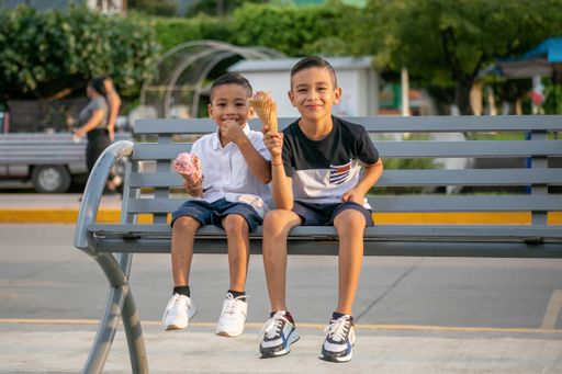 Two Hispanic cheerful kids sitting on a bench in a park and eating ice cream