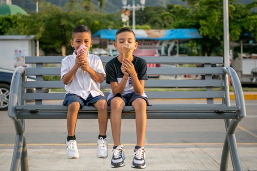 Two Hispanic cheerful kids sitting on a bench in a park and eating ice cream