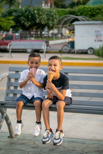 Two Hispanic cheerful kids sitting on a bench in a park and eating ice cream