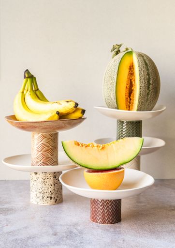 An artistic arrangement of bananas, melons, and grapefruit on decorative stands against a neutral background.