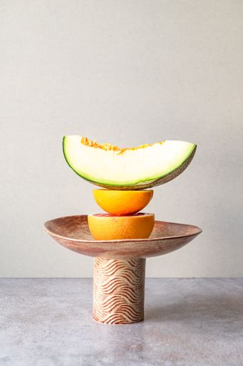 Artistic fruit arrangement featuring a melon slice, orange, and grapefruit on a decorative stand, set against a neutral background.