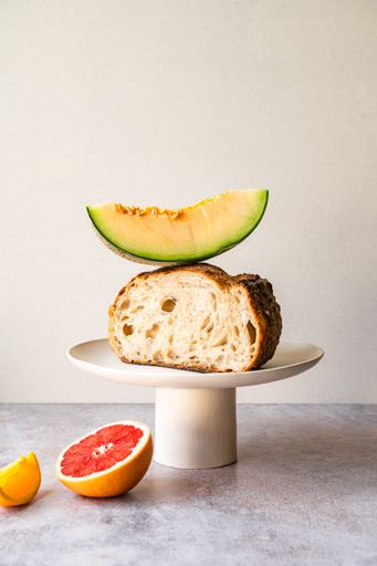 Artistic arrangement of bread, melon slice, and citrus fruits on a pedestal against a neutral background.
