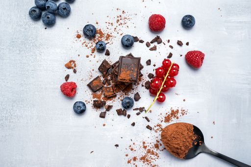 Flat lay of chocolate pieces, fresh blueberries, raspberries, red currants, and cocoa powder on a light textured background.