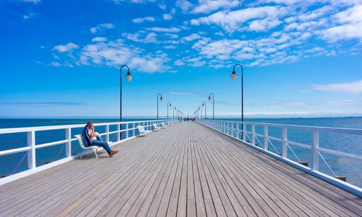 A long wooden pier with lamp posts and a person sitting on a bench at the seaside with a blue sky.