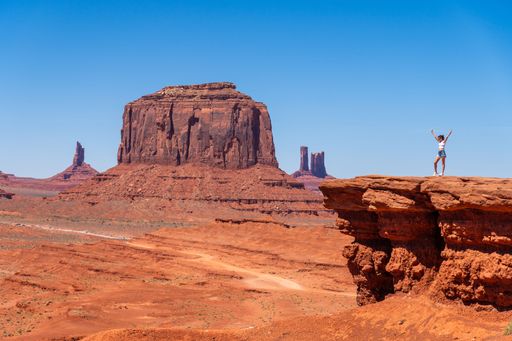 A person stands triumphantly on a rock ledge overlooking Monument Valley's iconic buttes under a clear blue sky.