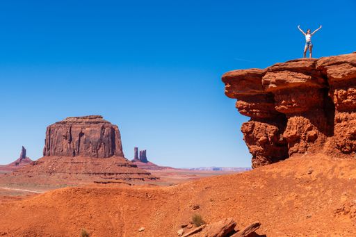 A person stands triumphantly on a rocky ledge overlooking Monument Valley's iconic red buttes under a clear blue sky.