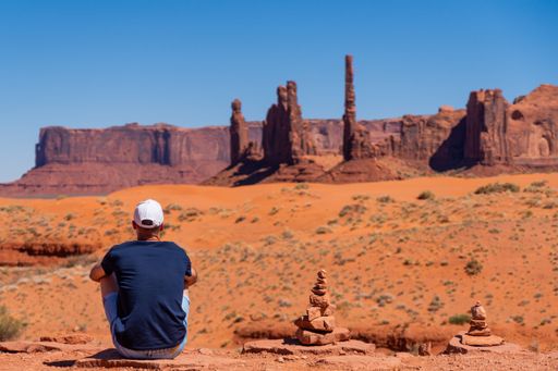 A man wearing a white cap sits in Monument Valley, Utah, USA, with rock formations and cairns under a clear blue sky.