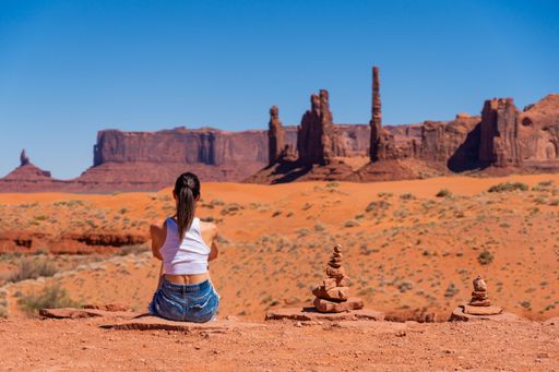A woman in casual attire sits on a desert landscape with rock formations under a clear blue sky, enjoying the view of Monument Valley.