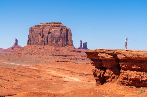 A woman joyfully stands on a cliff edge in Monument Valley, showcasing the stunning red rock formations under a clear blue sky.