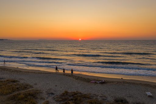 People enjoying a serene sunset on a sandy beach with gentle waves and a warm orange sky.