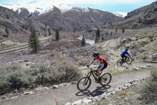 Two mountain bikers ride the Tahoe-Pyramid Trail with the Truckee River and Sierra Nevada peaks in the background.