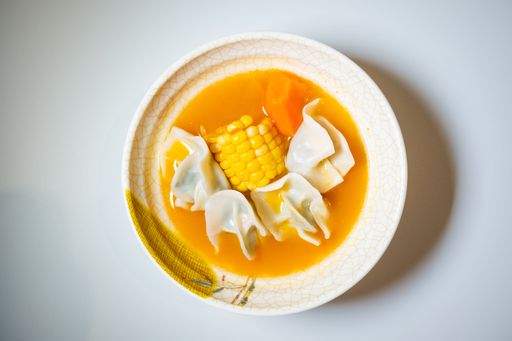 A top view of a bowl of soup with dumplings, corn, and carrots, served on a patterned plate.