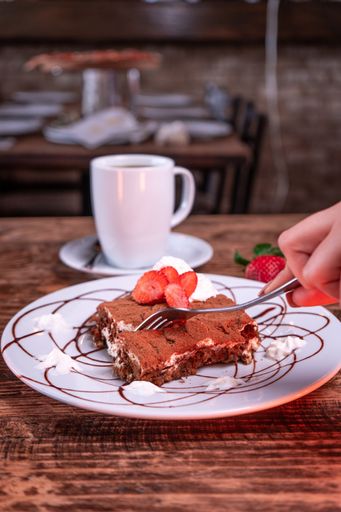 A person cutting a chocolate biscuit with strawberry next to a cup of coffee
